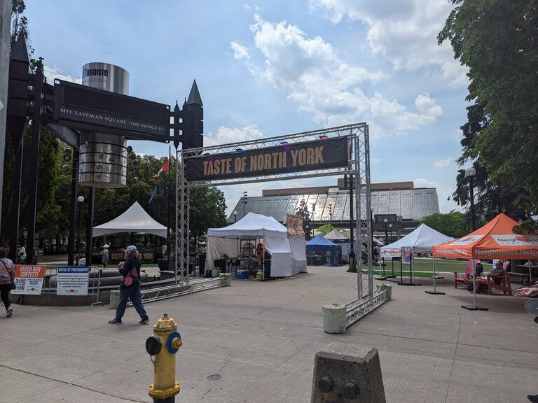 This is showing a picture from the festival which is still being set up. There are little pop-up tents, and a big overheard sign which reads 'Taste of North York'. To the left we can see the sign which is telling that this is also 'Mel Lastman Square'. Mel Lastman was the first Mayor of Toronto after it was almalgamated.